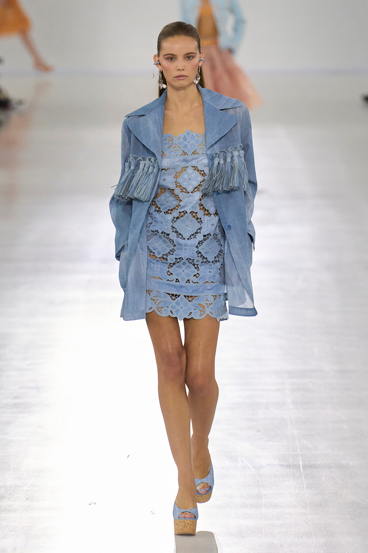 Runway model wearing a light-blue lace mini dress with a matching blue fringe-embellished jacket and platform wedges.