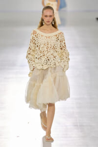Model walking a fashion show in a cream crochet-off-shoulder top and layered ivory tulle skirt, accessorized with statement earrings and white sandals.