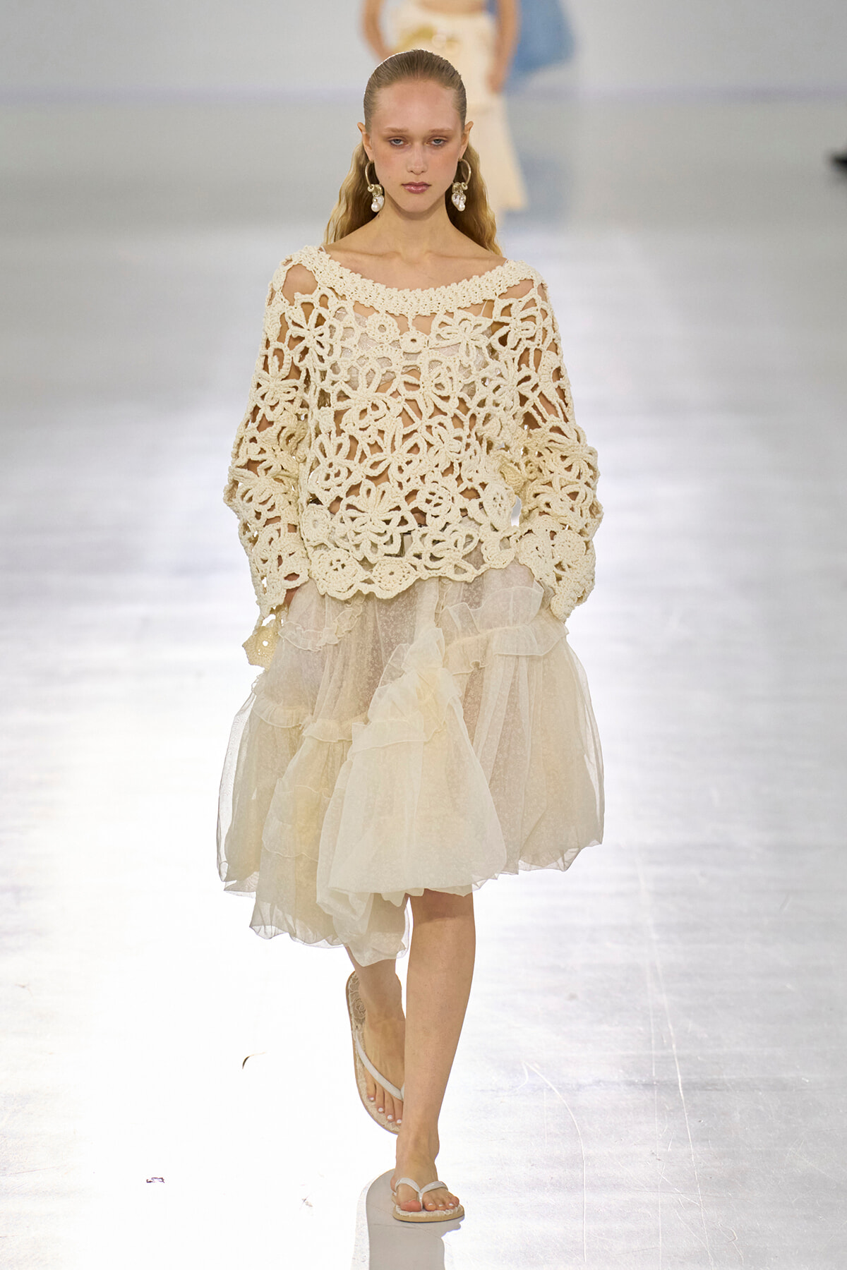 Model walking a fashion show in a cream crochet-off-shoulder top and layered ivory tulle skirt, accessorized with statement earrings and white sandals.