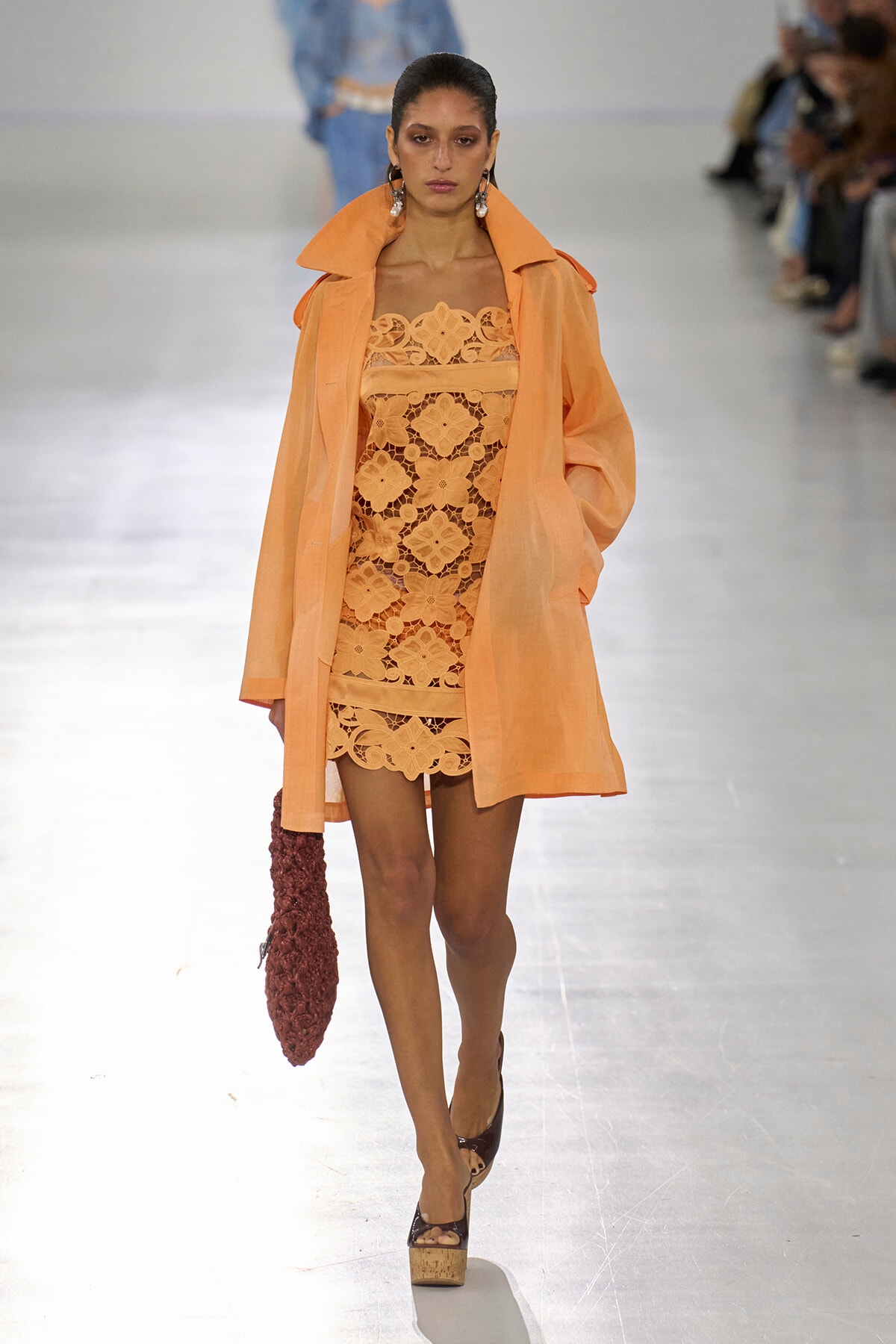 Model walking the runway in a peach lace mini dress and matching oversized orange trench, holding a burgundy knitted bag.