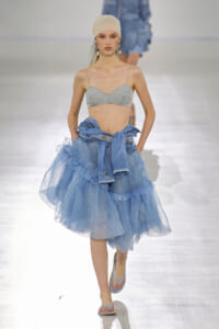 Model on a runway wearing a light blue lace bralette, a voluminous pale-blue tulle skirt, and a denim jacket tied around the waist with a headscarf and sandals.