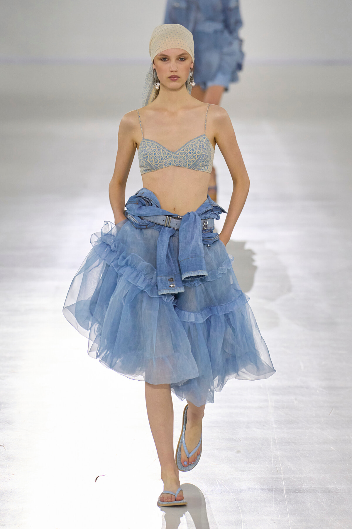 Model on a runway wearing a light blue lace bralette, a voluminous pale-blue tulle skirt, and a denim jacket tied around the waist with a headscarf and sandals.