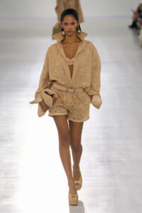 Model walks the runway in a beige lace shorts set with a matching belt, open blouse, and nude wedges, carrying a woven clutch.