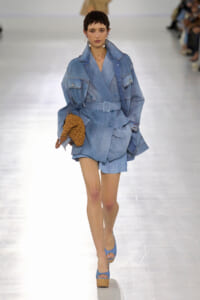 Runway model in a light-blue denim trench with belt, matching shorts, blue platform sandals, and a brown woven bag in hand.