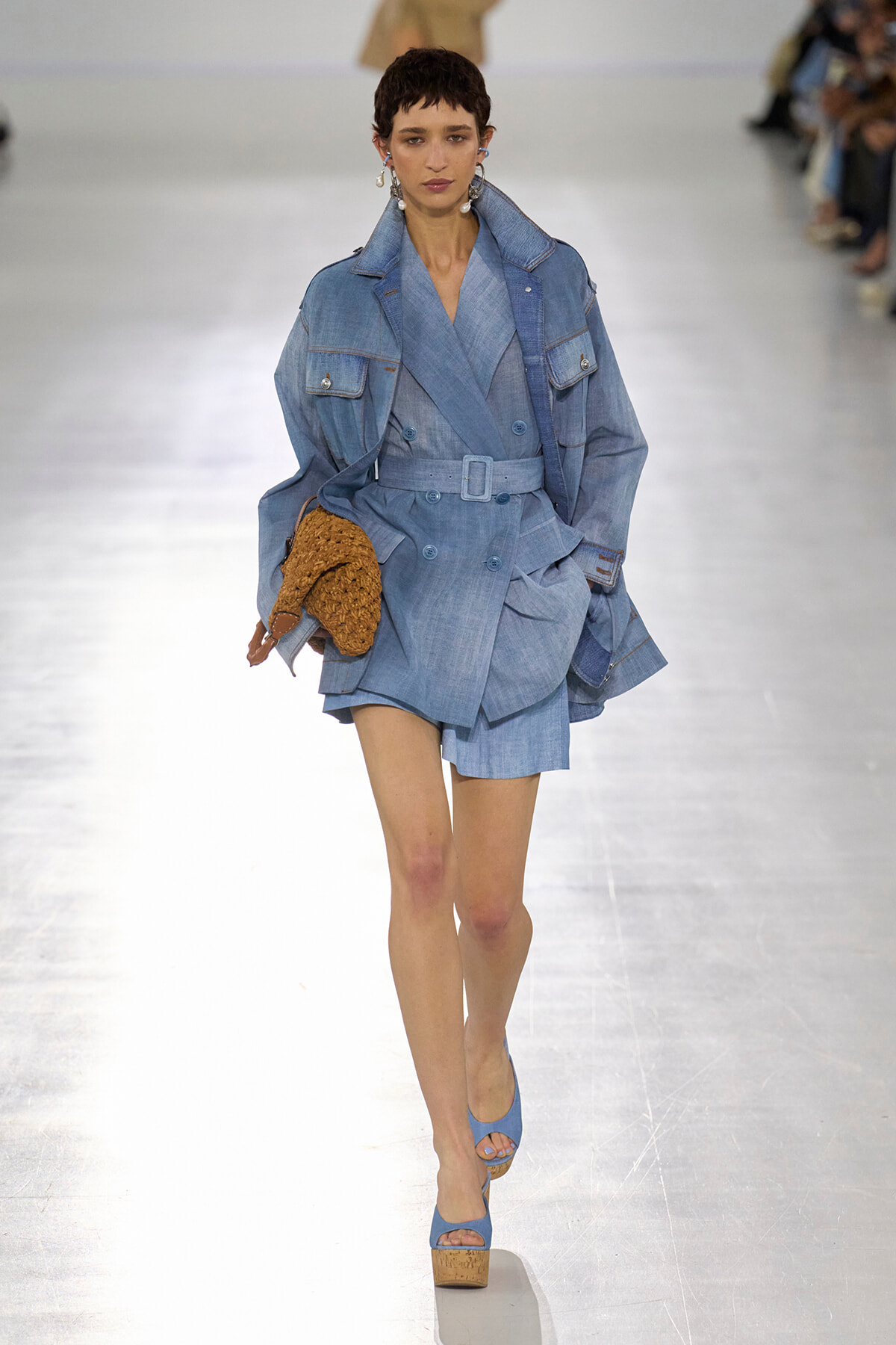 Runway model in a light-blue denim trench with belt, matching shorts, blue platform sandals, and a brown woven bag in hand.