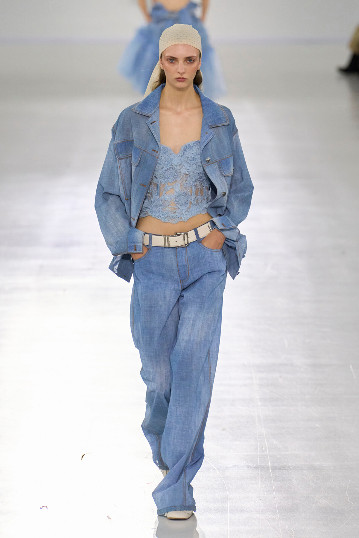 Model on a runway wearing a light blue denim suit: oversized jacket, high-waisted jeans, lace crop top, white belt, and beige beanie.