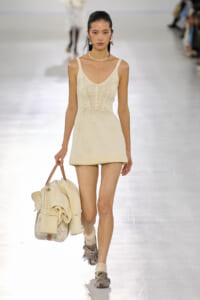Model walks a fashion runway in a cream knit mini dress, matching knit tote bag slung in her left hand and pearl jewelry.