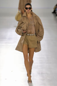 Model walks a fashion runway in a tan trench, matching shorts, lace top, and belt, wearing oversized sunglasses and carrying a clutch.