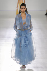 Model walking the runway in a light blue sheer dress with a lace bralette and a sparkly belt on a fashion show catwalk.