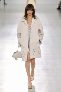Model on a runway wearing a pale beige coat with lace appliqué and a small white handbag in front of a bright runway background.
