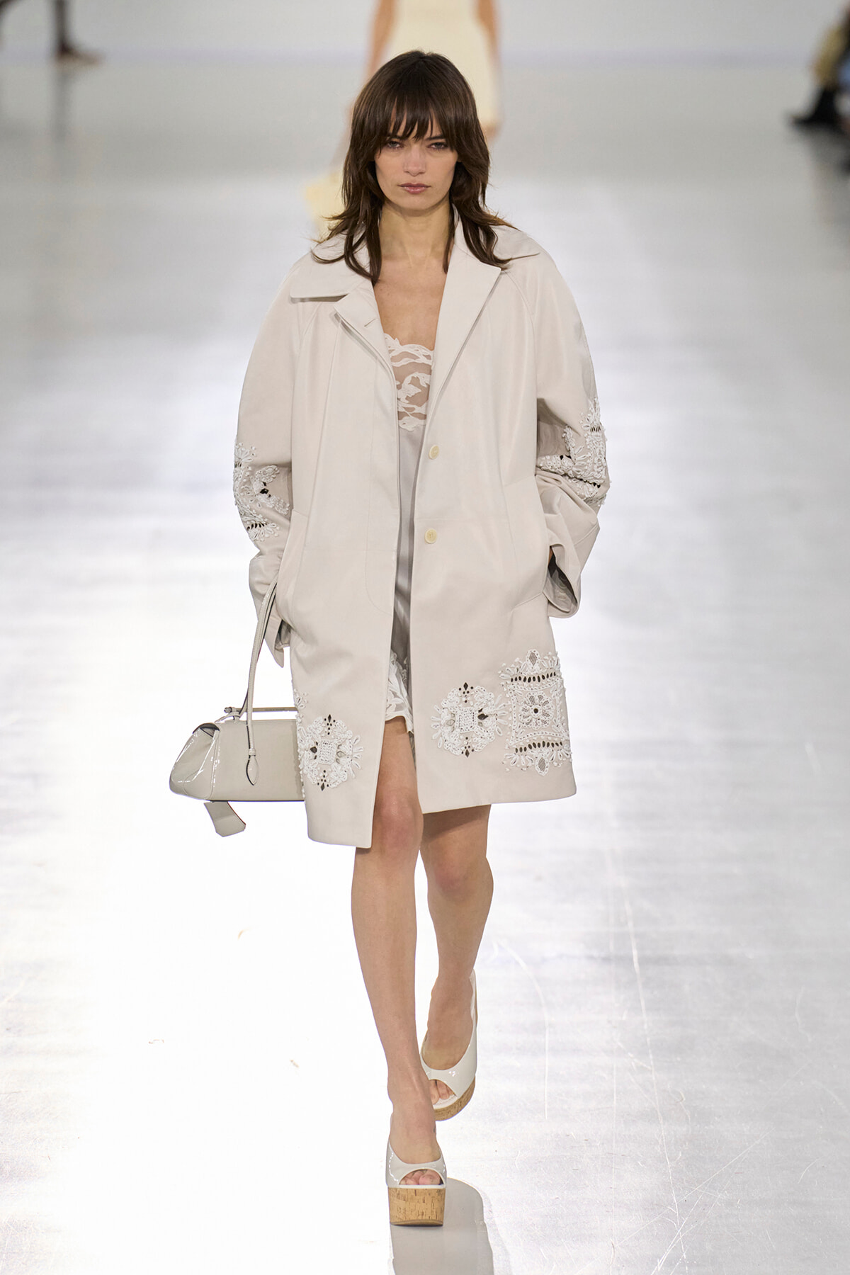 Model on a runway wearing a pale beige coat with lace appliqué and a small white handbag in front of a bright runway background.