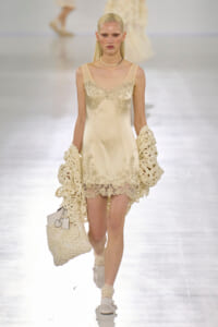 Model walks the runway in an ivory satin slip dress with lace trim, paired with crochet cream gloves and a matching crocheted bag. Pearl choker."
