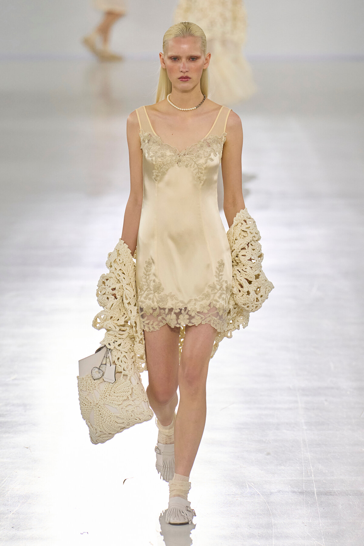 Model walks the runway in an ivory satin slip dress with lace trim, paired with crochet cream gloves and a matching crocheted bag. Pearl choker."