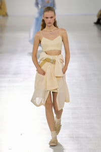 Model walks a runway wearing a cream crop top and high-waisted wrap skirt with a jeweled belt and pearls necklace.