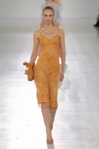 Model walks the runway in an orange lace sheer midi dress with thin straps, holding a matching clutch and wearing a pearl necklace.