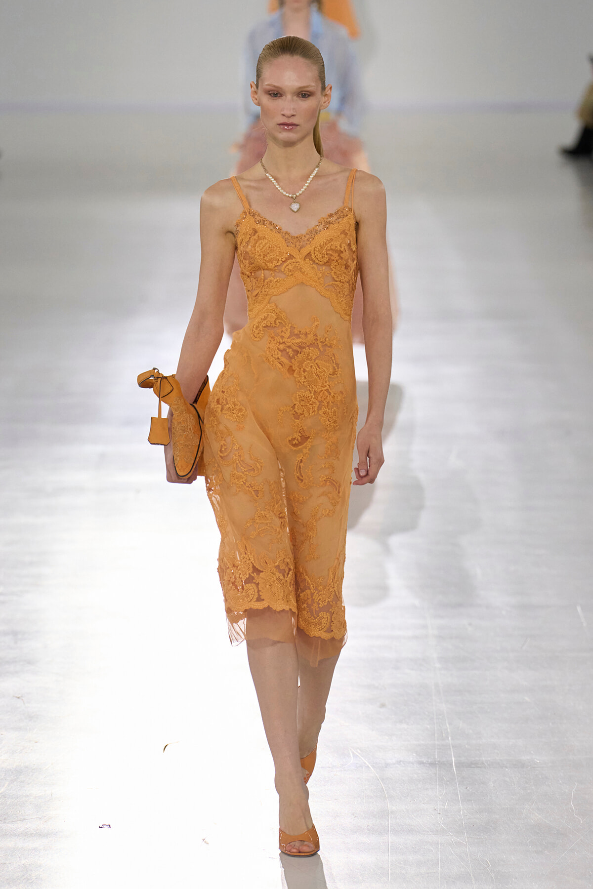 Model walks the runway in an orange lace sheer midi dress with thin straps, holding a matching clutch and wearing a pearl necklace.