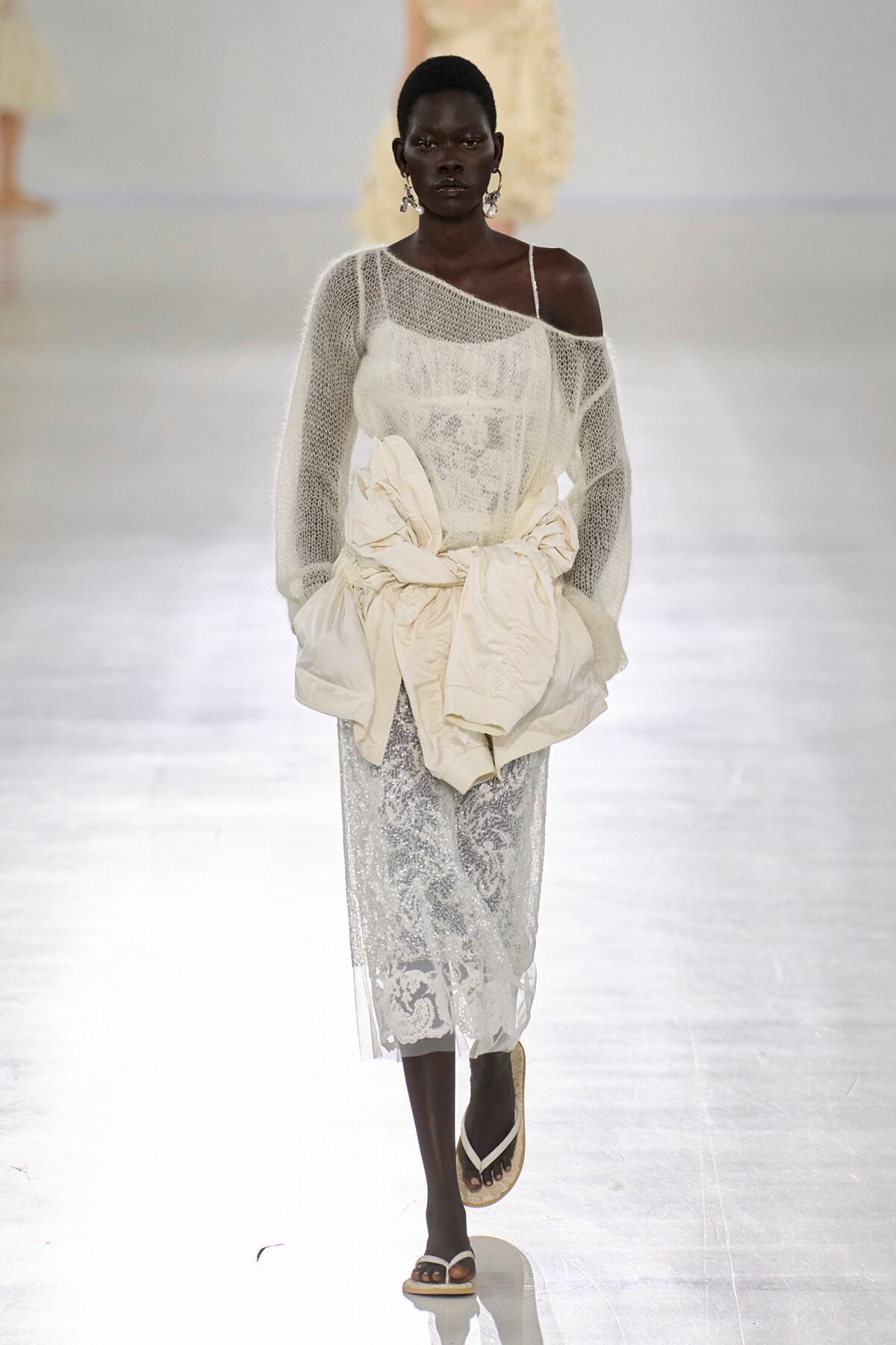 Model walks a runway in a white lace midi dress with a sheer knit top, an oversized cream bow at the waist, and dangling earrings. She wears thong sandals on a glossy catwalk.