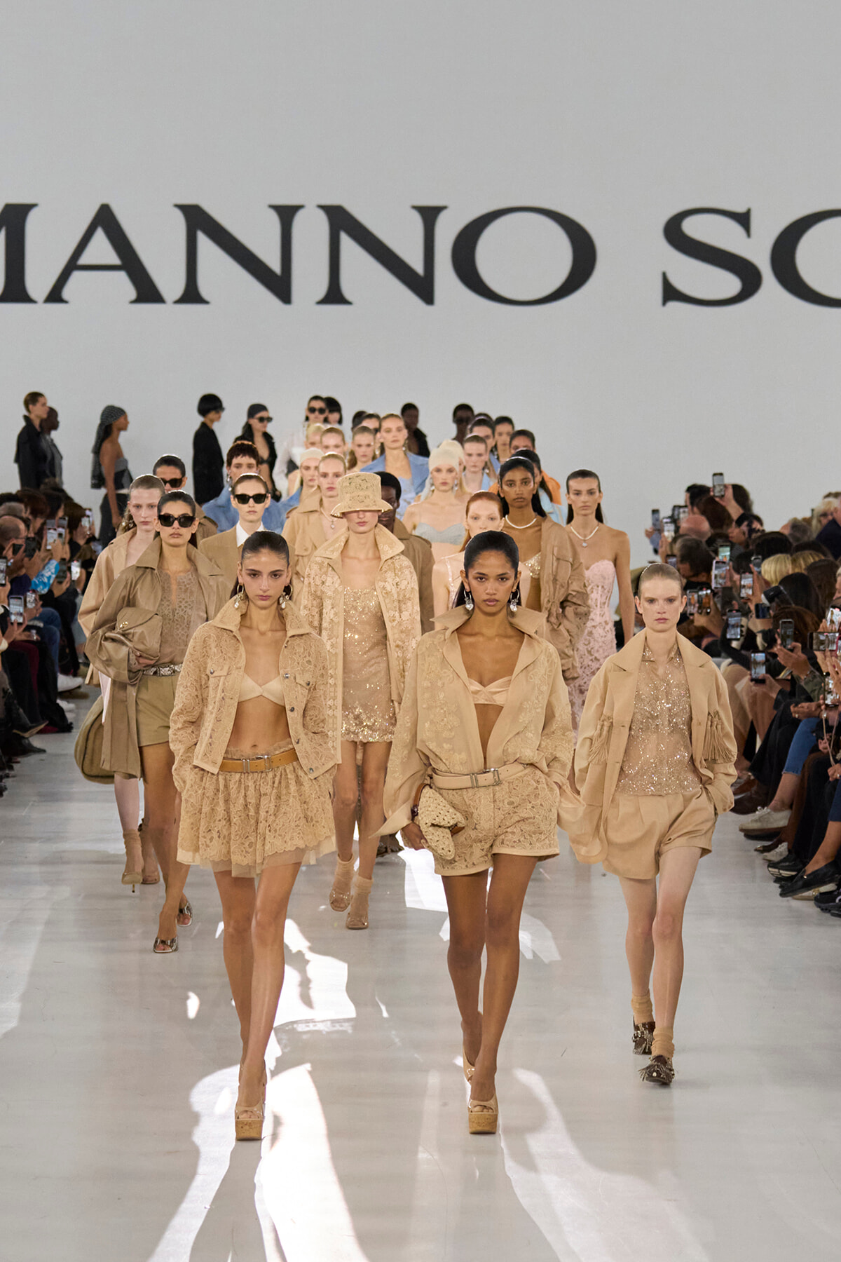 Models in beige outfits walk a fashion runway, audience on both sides filming and watching the show.