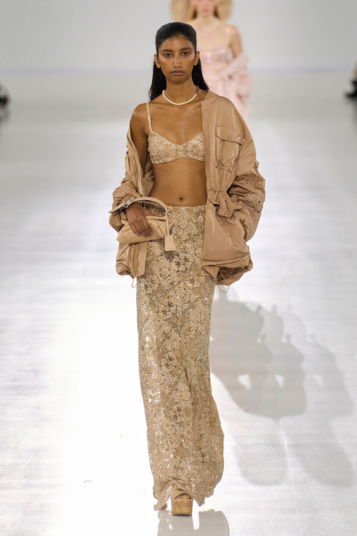Model on a runway wearing a beige sequin two-piece dress with a matching oversized jacket and pearl necklace.