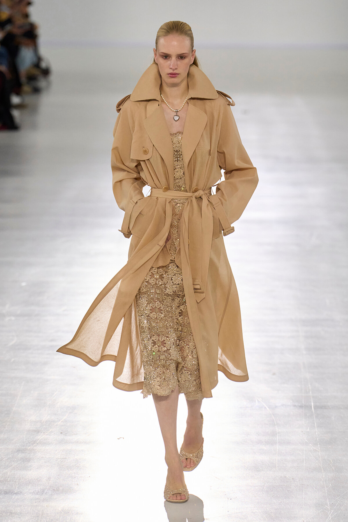 Runway model wearing a camel trench coat over a gold sequined midi dress, belt tied at the waist, with pearl necklace and beige sandals on the catwalk.