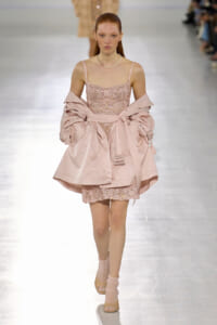 Model walking the runway in a pale pink sequin mini dress with thin straps, paired with a matching oversized pink coat tied at the waist. A pearl necklace completes the look.