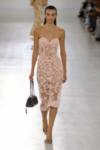 Model on a runway wearing a blush pink, lace strapless midi dress with a pearl necklace and small black handbag.