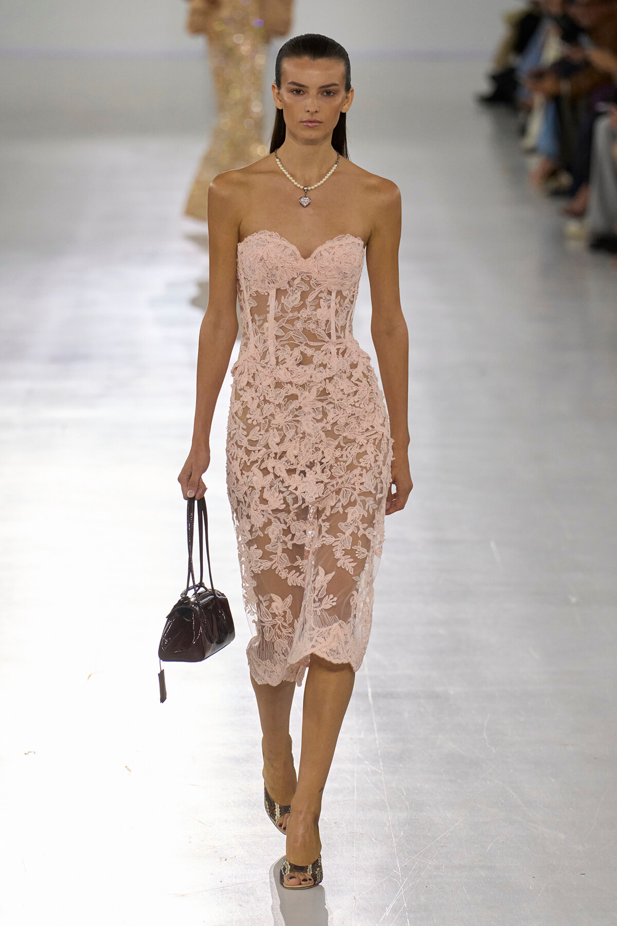 Model on a runway wearing a blush pink, lace strapless midi dress with a pearl necklace and small black handbag.