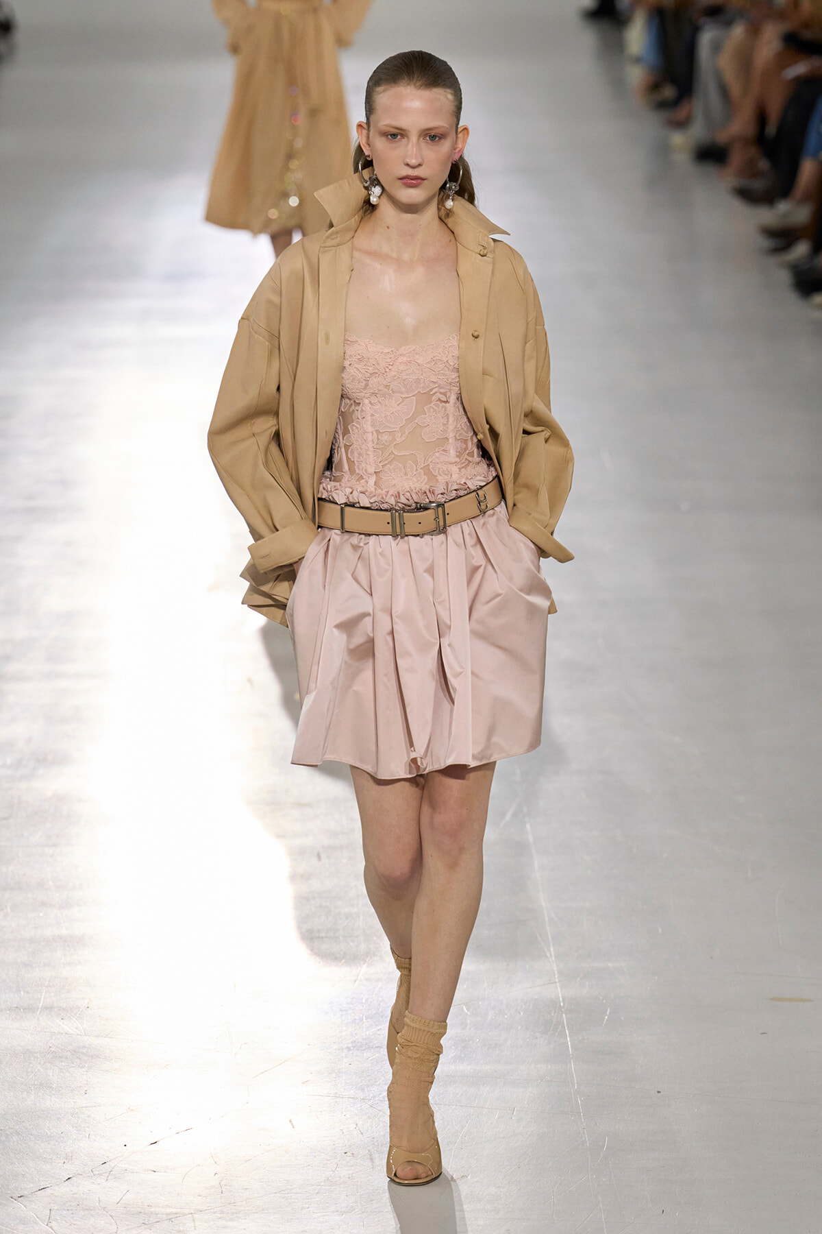 Model walks the runway in a beige oversized jacket over a pink lace bodysuit and satin pink pleated skirt with a tan belt.