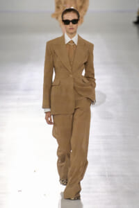 Model in a tan tailored pantsuit with white shirt and pearl necklace walking the runway, wearing dark sunglasses on glare-lit catwalk.