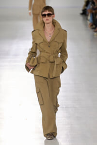 Model walking the runway in a tan safari-inspired belted utility suit with oversized collar, sunglasses, and a pearl necklace.
