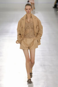 Model walks the runway in a beige coordinated outfit: fringe jacket over a sequined top and matching shorts, with embellished boots.