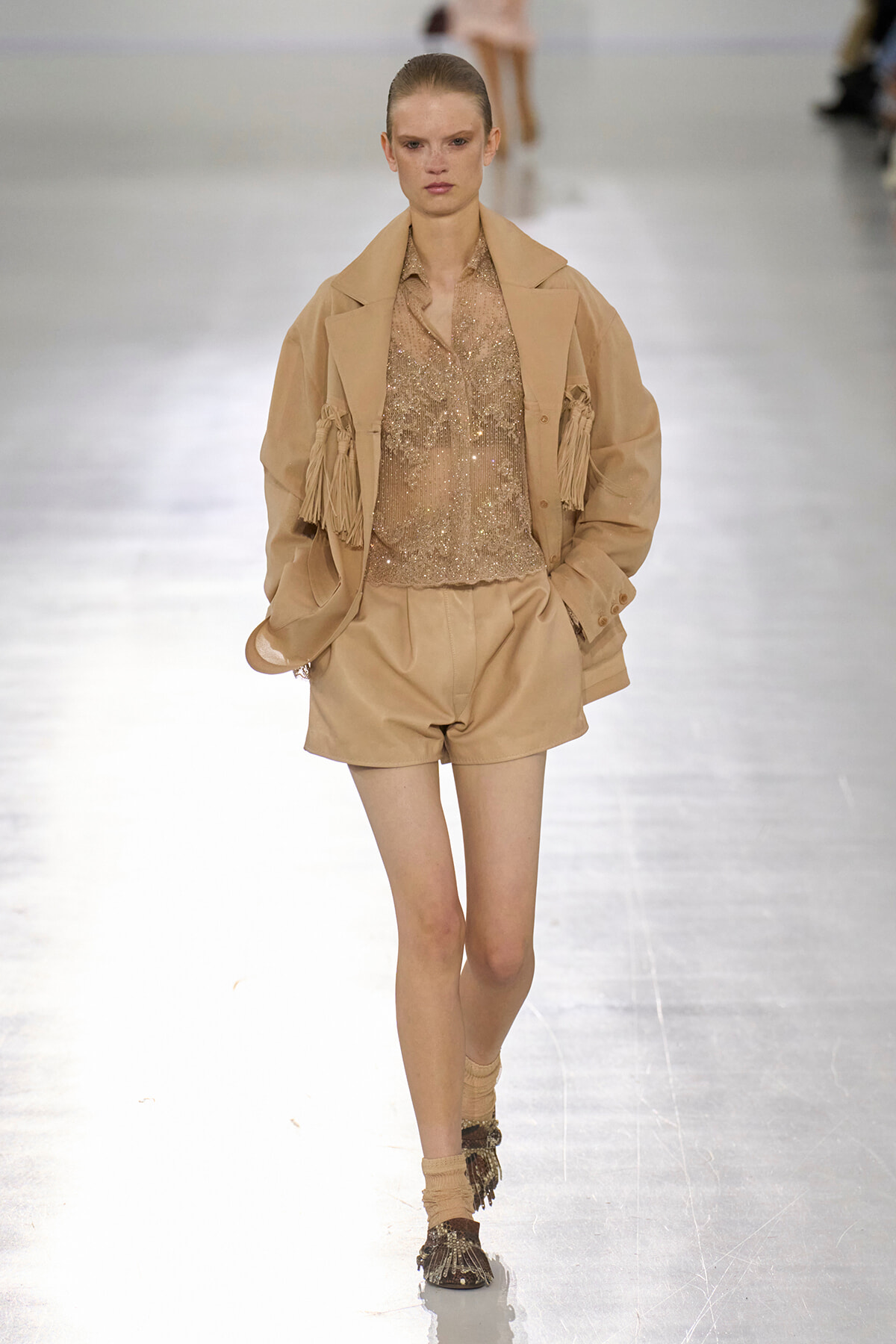 Model walks the runway in a beige coordinated outfit: fringe jacket over a sequined top and matching shorts, with embellished boots.