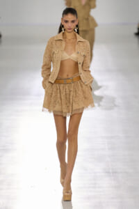 Model on a runway wearing a beige lace jacket over a matching bralette and a short lace skirt with a tan belt and platform sandals.
