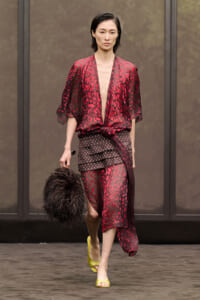 Model walking a runway in a red leopard-print sheer blouse tied at the waist, with a layered brown polka-dot skirt and lime-green heels, carrying a black fluffy handbag.