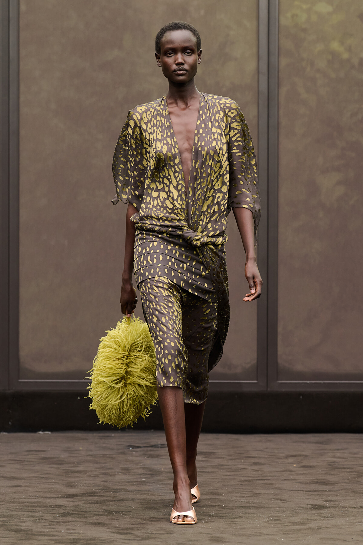 Model walking a runway in a brown and olive animal-print set with a knotted waist, carrying a lime-green shaggy handbag and wearing pale heels.