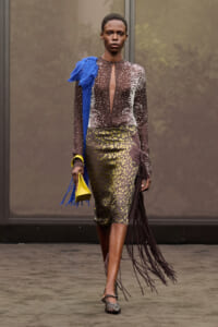 Model on a runway wearing a sheer brown, dotted top with a cutout, an olive animal-print midi skirt, a blue fringe scarf, and a bright yellow clutch and fringe skirt detail on the side.