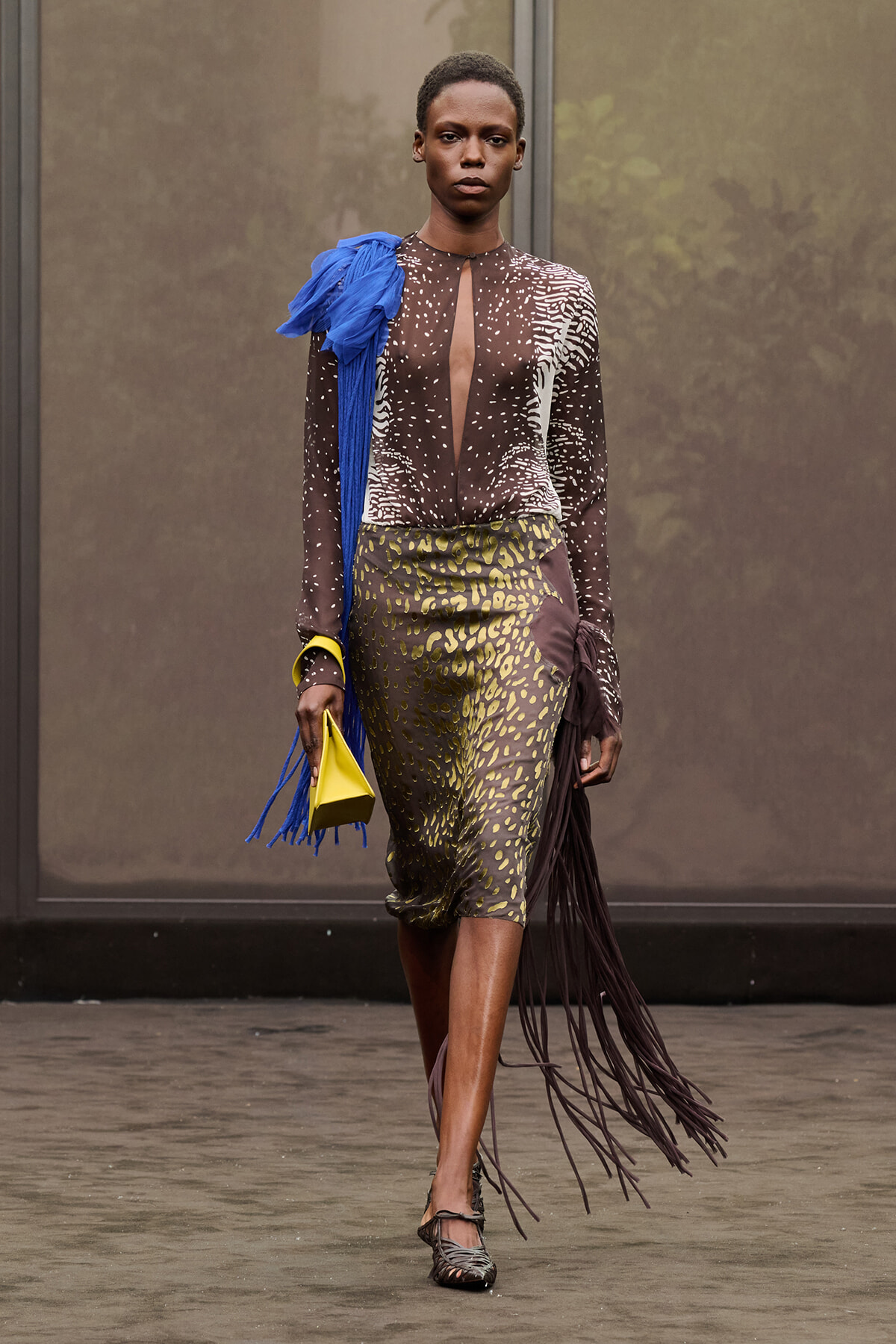 Model on a runway wearing a sheer brown, dotted top with a cutout, an olive animal-print midi skirt, a blue fringe scarf, and a bright yellow clutch and fringe skirt detail on the side.
