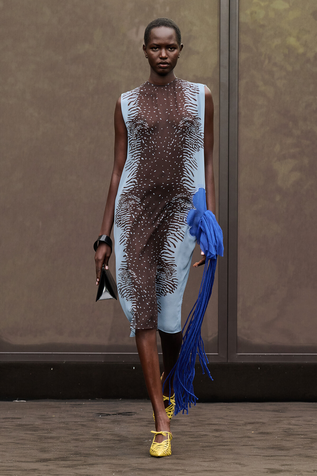 Model walking a runway in a sheer black-and-blue patterned dress with light blue panels, holding a blue fringe accessory and a small black clutch, wearing yellow sandals.