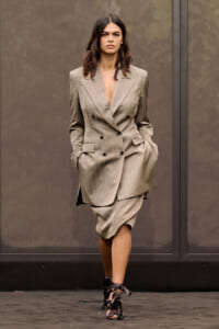 Female model walking a runway in a taupe oversized double-breasted blazer with a matching knee-length skirt, hands in pockets.