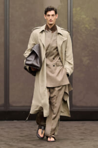 Male model walking a runway in a beige trench coat over a layered taupe suit, carrying a brown leather bag.
