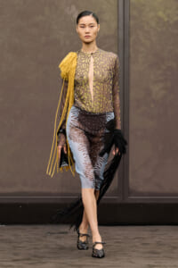 Model walking a runway in a sheer animal-print blouse with mustard fringe over blue-black patterned midi pants and black woven heels.