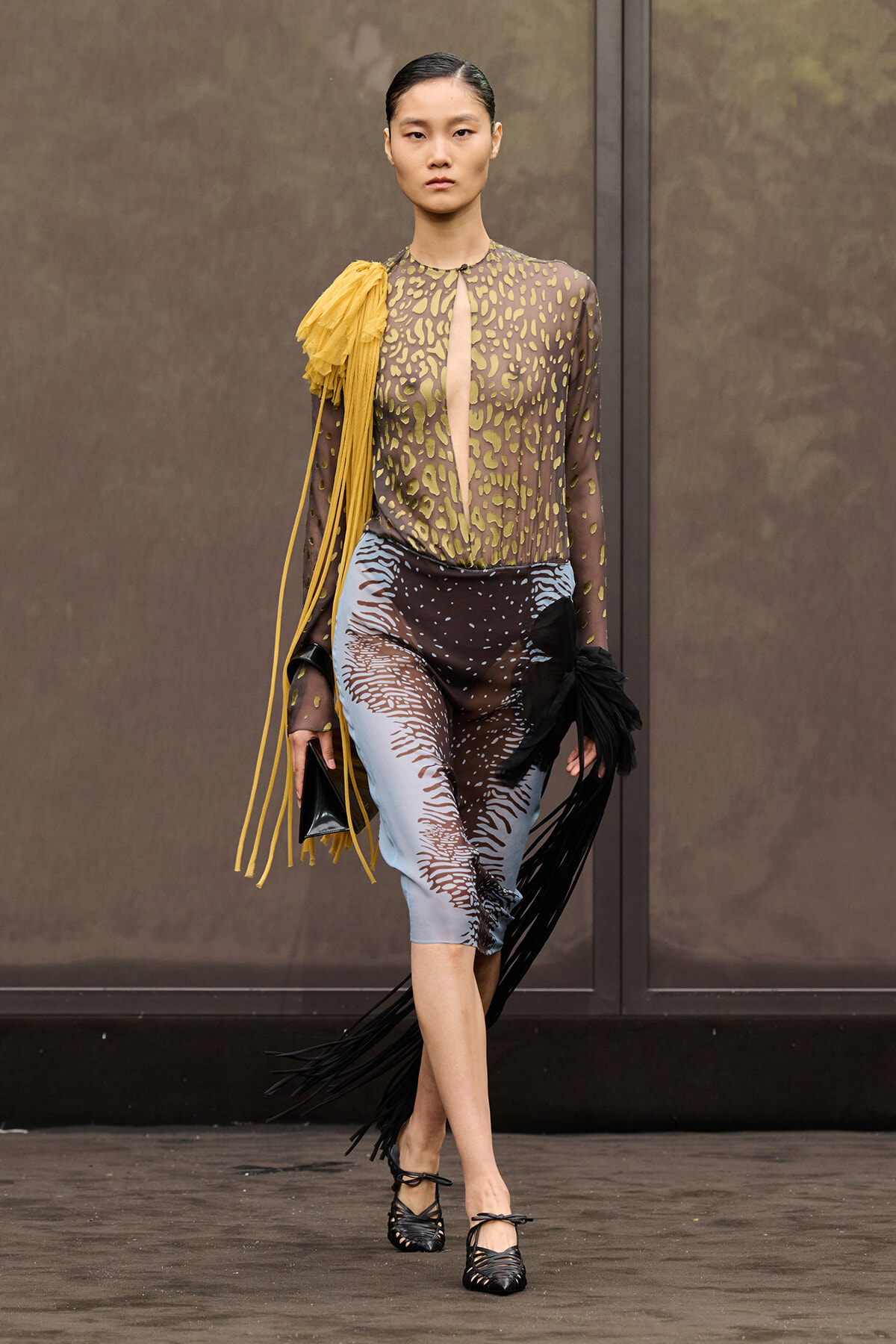 Model walking a runway in a sheer animal-print blouse with mustard fringe over blue-black patterned midi pants and black woven heels.