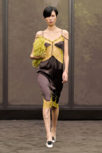 Model walks a runway in a black satin dress with yellow lace panels and spaghetti straps, holding a lime-green feathered clutch.