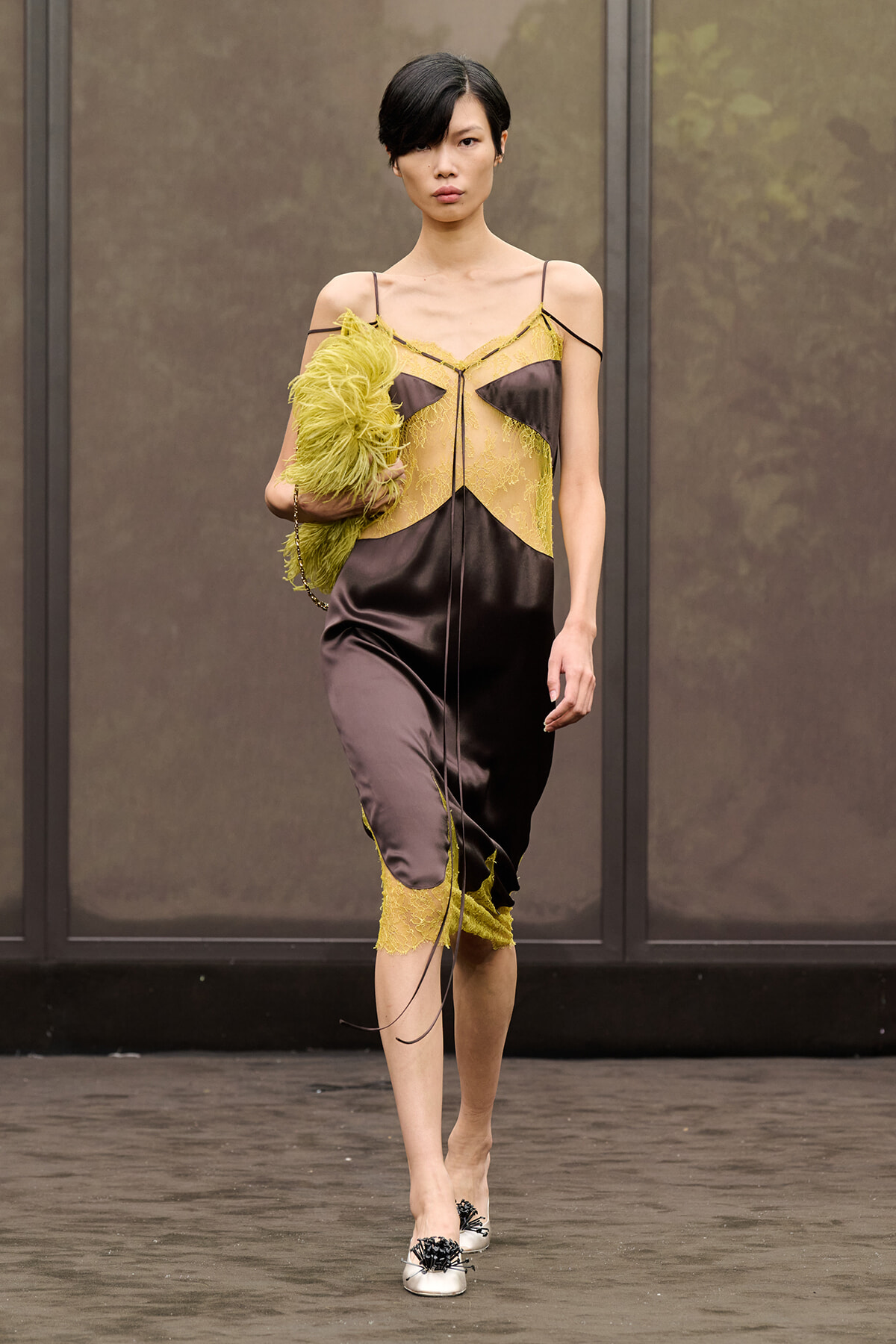 Model walks a runway in a black satin dress with yellow lace panels and spaghetti straps, holding a lime-green feathered clutch.