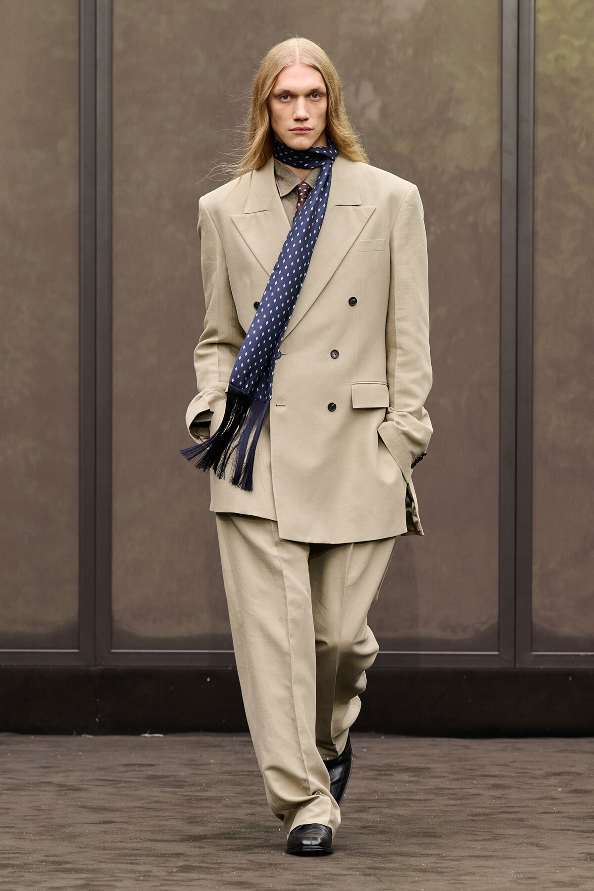Male model walks the runway in a beige double-breasted suit with a navy patterned scarf and fringe tassels.