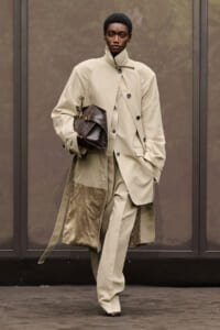 Model walking a runway in a beige layered trench coat over a matching suit, holding a dark brown woven handbag under the arm.