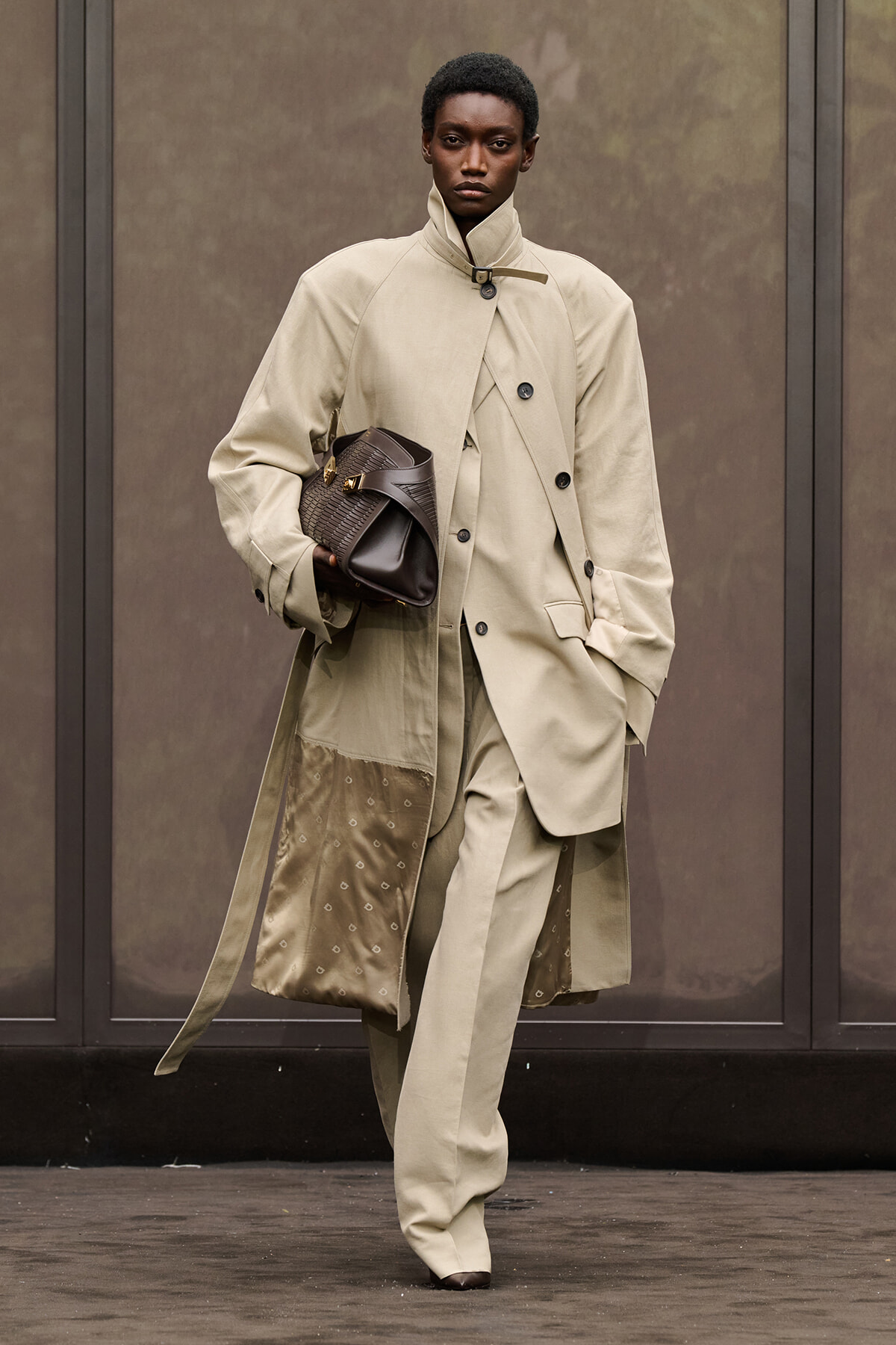 Model walking a runway in a beige layered trench coat over a matching suit, holding a dark brown woven handbag under the arm.