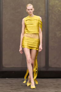 Model on a runway wearing a mustard yellow layered dress with a sheer lace midriff and long ribbon accents.