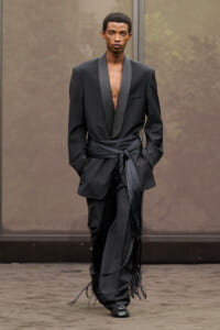 Male model walking a runway in a black satin-scarf belted suit with deep V-neck and fringe detailing at the cuffs and hem.