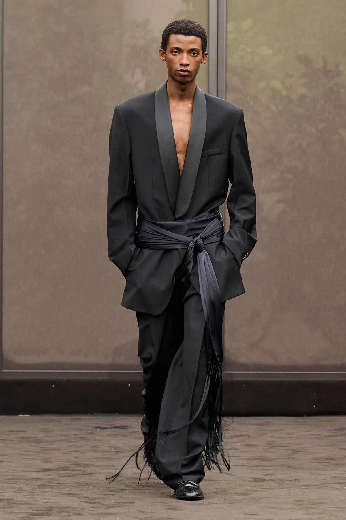 Male model walking a runway in a black satin-scarf belted suit with deep V-neck and fringe detailing at the cuffs and hem.
